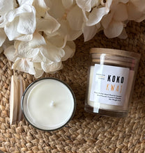 Load image into Gallery viewer, Two candles in glass jars with wooden lids sit on a woven surface with white flowers-they're labeled with the scent of Koko Kwaj.
