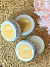 Load image into Gallery viewer, Three travel candle tins with a yellow label in the Lauderdale scent on a woven surface with flowers.
