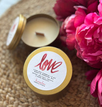 Load image into Gallery viewer, Candle in a gold tin with 'love' label next to pink flowers on a woven surface
