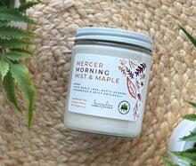 Load image into Gallery viewer, Glass candle jar labeled with scent &#39;Mercer Morning Mist &amp; Maple&#39; on a woven surface with plants around.
