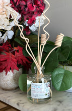 Load image into Gallery viewer, A reed diffuser bottle with a clear glass jar, clear liquid, and natural curly wood reeds, placed on a marble surface with flowers and green leaves in the background. The scent label reads 'MIDNIGHT SNOW ON GRAND'.
