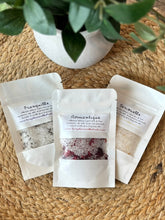 Load image into Gallery viewer, Three small white packages filled with bath salts on a woven surface, with a plant in the background.
