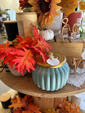 Decorative pumpkin-shaped candle with a gold top and blue base in a store setting with autumn leaves and other decorations.