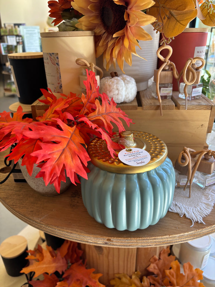 Decorative pumpkin-shaped candle with a gold top and blue base in a store setting with autumn leaves and other decorations.