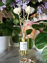 Load image into Gallery viewer, A reed diffuser bottle with white tulip-shaped reeds and a label showing the candle scent, Pure Maui, sits on a marble surface surrounded by potted plants and shells.
