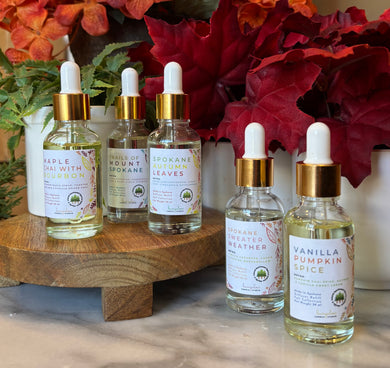 Five bottles of fragrance oils in winter scents with dropper lid, grouped on a wooden stand with autumn leaves in the background.