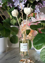 Load image into Gallery viewer, A reed diffuser bottle with white tulip-shaped reeds and a label showing the candle scent, Rock Away, sits on a marble surface surrounded by potted plants and shells.
