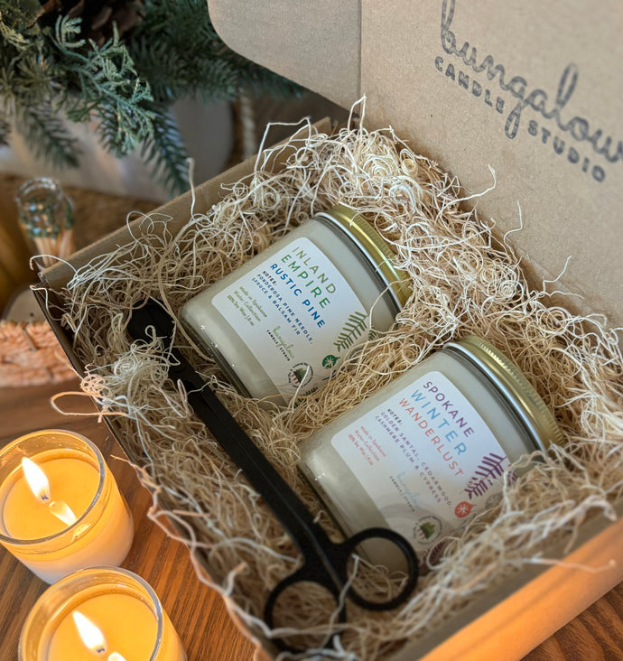 Two cylindrical candles in clear glass jars, set in a rustic-looking box with holiday decorations, including a wick trimmer. The candles are labeled with different scents 