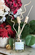 Load image into Gallery viewer, A clear glass reed diffuser bottle with a white scent label reading 'INLAND EMPIRE RUSTIC PINE' and natural wood reeds with spiral and wavy tops, styled on a marble surface with red and white flowers and green leaves in the background.
