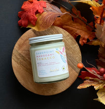 Load image into Gallery viewer, Candle jar labeled &#39;Sandpoint Mahogany Tobacco&#39; on a wooden board with fall leaves.
