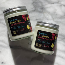 Load image into Gallery viewer, 8oz candle labeled &#39;Snow Country Coeur d&#39;Alene&#39; with silver lid on marble surface
