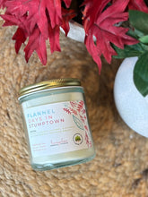 Load image into Gallery viewer, Candle labeled 'Flannel Days in Stumptown' with a red decorative plant in the background on a woven mat
