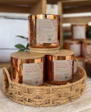 Three copper-colored candles labeled 'Spokane Sweater Weather' soy candles in a woven basket.