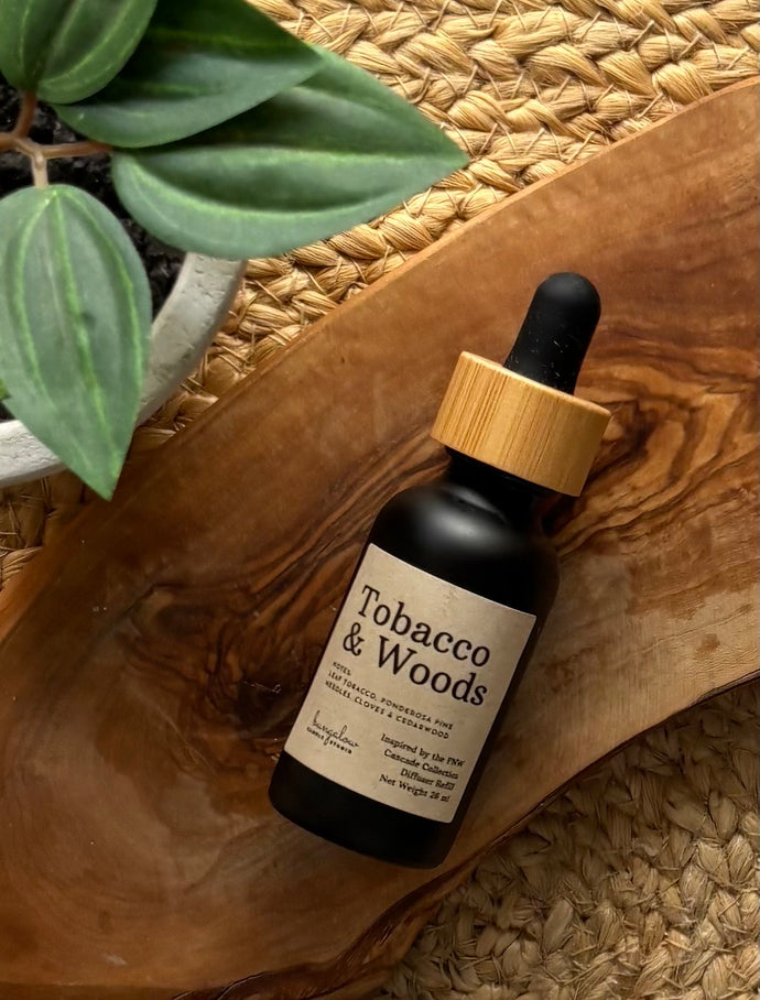 Black dropper bottle of Cascade car diffuser in Tobacco & Woods scent on a wooden surface with a plant in the background