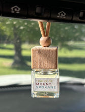 Load image into Gallery viewer, A car diffuser in the Trails of Mount Spokane scent from the Fall Candle Scent Collection hangs from the car rearview mirror.

