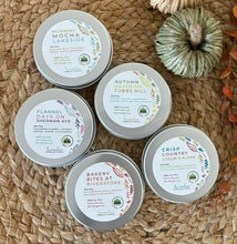 Load image into Gallery viewer, Five silver metal tin candles with labels indicating various fall scents, arranged on a woven surface with decorative pumpkins and leaves in the background.
