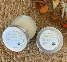 Load image into Gallery viewer, Two silver metal tin travel candles in the scent, &quot;Autumn Haven on Tubbs Hill,&quot; arranged open and closed on a woven surface with decorative fall leaves in the background.
