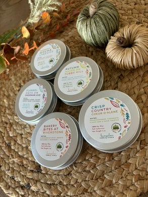 Five silver metal tin candles with labels indicating various fall scents, arranged on a woven surface with decorative pumpkins and leaves in the background.