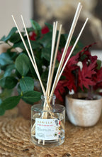Load image into Gallery viewer, Glass reed diffuser jar in the Vanilla Peppermint scent with wood reeds sitting on a table with green and red plants in background.
