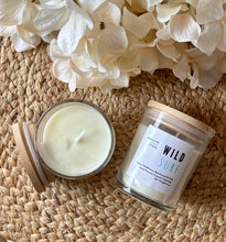 Load image into Gallery viewer, Two candles in glass jars with wooden lids sit on a woven surface with white flowers-they're labeled with the scent of Wild Surf.
