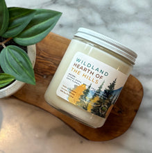 Load image into Gallery viewer, Candle jar labeled &#39;Wildland Hearth of the Hills&#39; on a wooden coaster with a plant in the background.
