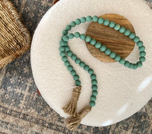 Load image into Gallery viewer, A string of sage green contentment beads made from Mango Wood draped on foot rest for home decor.
