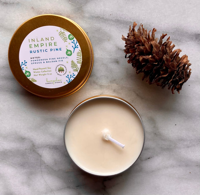 A gold travel tin candle labeled in the scent of Inland Empire Rustic Pine presented open with a pine cone added for decoration.