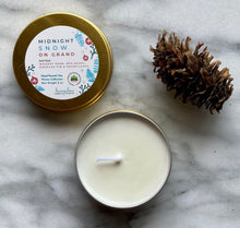 Load image into Gallery viewer, A gold travel tin candle labeled in the scent of Midnight Snow on Grand presented open with a pine cone added for decoration.
