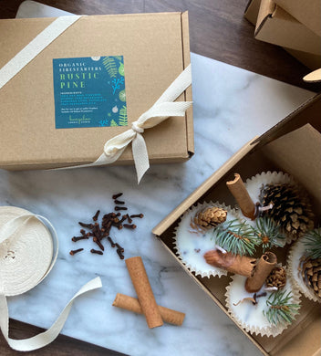 A gift box labeled 'Organic Fire Starters' with a rustic pine design, containing 4 organic fire starters with pinecone, cloves, and cinnamon sticks visible. The box is presented with a bow and appears to be made of recyclable material.