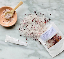 Load image into Gallery viewer, An overhead shot of a bath salt product with its packaging. The bath salt is scattered on a surface, coming from a white Kraft paper pouch that it is packaged in.
