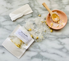 Load image into Gallery viewer, An overhead shot of a bath salt product with its packaging. The bath salt is scattered on a surface, coming from a white Kraft paper pouch that it is packaged in.
