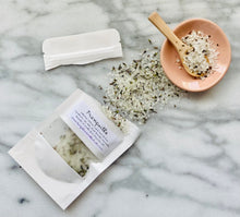 Load image into Gallery viewer, An overhead shot of a bath salt product with its packaging. The bath salt is scattered on a surface, coming from a white Kraft paper pouch that it is packaged in.
