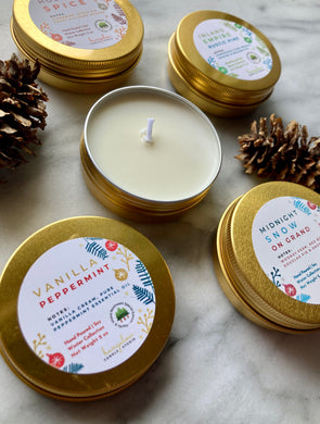 A set of travel tin candles with holiday and winter scents, including vanilla peppermint and rustic pine. The candles are presented in gold metal tins with decorative labels, and some are surrounded by pine cones.