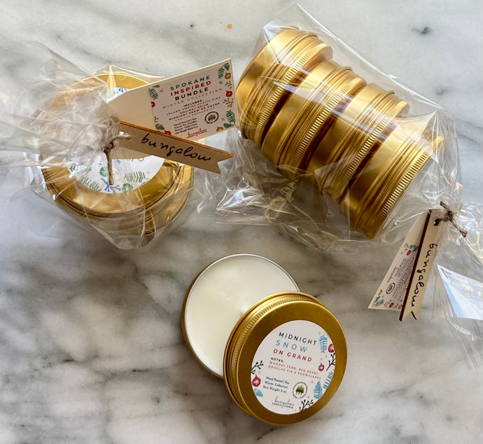 A set of travel tin candles with holiday and winter scents, including vanilla peppermint and rustic pine. The candles are presented in gold metal tins with decorative labels.