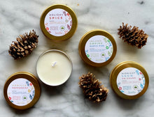 Load image into Gallery viewer, A set of travel tin candles with holiday and winter scents, including vanilla peppermint and rustic pine. The candles are presented in gold metal tins with decorative labels, and some are surrounded by pine cones.
