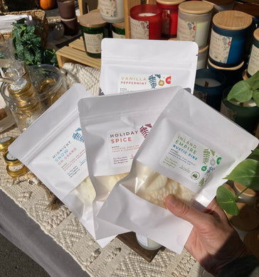 4 Packages of soy wax melts in white pouches with a variety of holiday and winter scents held up in front of a display table.